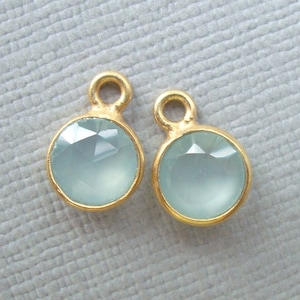 May include: Two gold-toned charms with light blue gemstone accents. Each charm has a round gemstone and a small loop at the top.