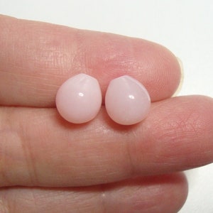 May include: Two pink teardrop-shaped beads, likely made of shell or pearl, are held between two fingers.