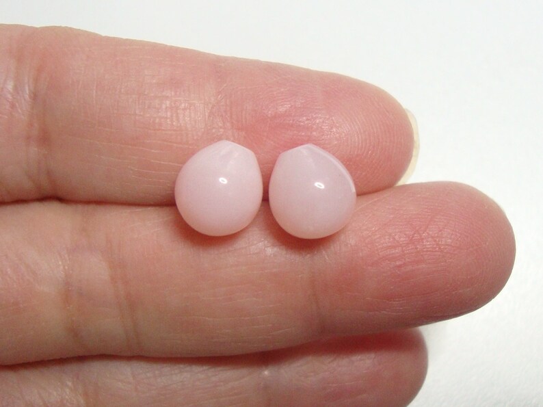May include: Two pink teardrop-shaped beads, likely made of shell or pearl, are held between two fingers.