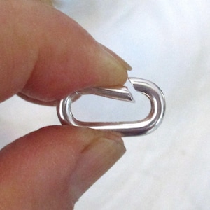 May include: A small, shiny, silver-colored oval carabiner clip. The clip is held open, showing the opening mechanism. The background is a soft, out-of-focus white.