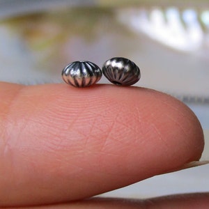 May include: Two silver-toned beads with a textured, scalloped design. The beads are small and round, and they are shown on a finger.