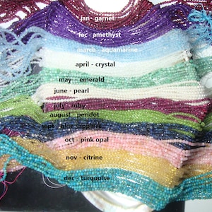 May include: A collection of gemstone beads, arranged in rows by month, with the corresponding birthstone name labeled below each row. The beads are all small and round, and they are strung on a thin thread. The colors of the beads are vibrant and varied, and they include red, purple, blue, green, yellow, and white.