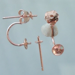 May include: A pair of rose gold earring findings with a curved post and a flat back. The findings are designed to hold a pearl or other gemstone.