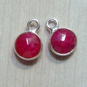 Op de afbeelding: Twee zilveren bedels met een ronde, felrode edelsteen in het midden. De bedels hebben een kleine lus aan de bovenkant om aan een ketting of armband te bevestigen.