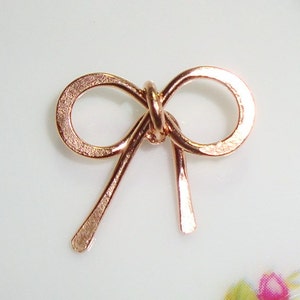 May include: A small, rose gold-colored metal bow charm. The bow is made of a single piece of wire that is bent into a bow shape. The bow is smooth and shiny.