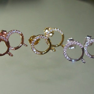 May include: Three pairs of earring findings with a round, open circle design. Each pair is a different metal color: rose gold, gold, and silver. Each circle is accented with small, clear rhinestones.