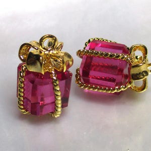 May include: Two gold-tone gift box charms with pink crystal accents. The charms feature a bow and rope detail.
