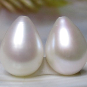 May include: Two pearlescent white teardrop-shaped pearls, each with a smooth, glossy surface, are displayed side-by-side on a white background.