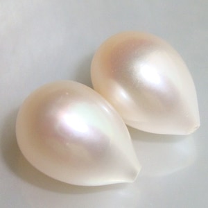 May include: Two pearlescent, teardrop-shaped beads. The beads have a creamy white color with subtle iridescent highlights. The smooth surface reflects light, creating a soft, glowing effect. The beads are likely for jewelry making or crafting.