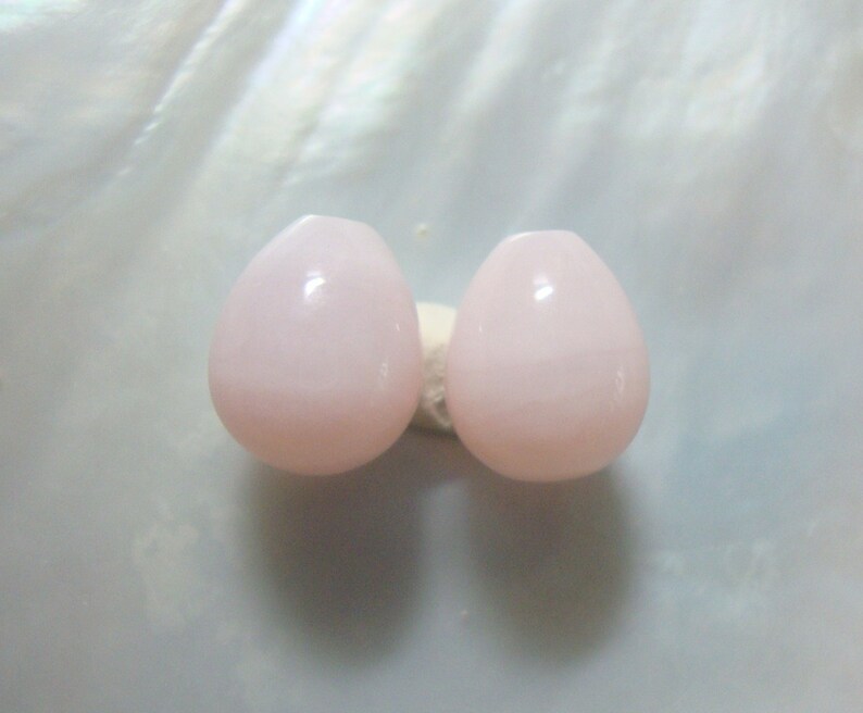 Puede incluir: Dos cuentas de color rosa en forma de l&aacute;grima con un acabado liso y brillante.