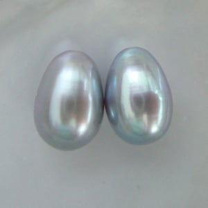 May include: Two iridescent, oval-shaped pearls with a smooth, reflective surface. The pearls have a soft, silvery-blue hue and a subtle sheen, suggesting a high-quality finish. The background is a neutral, light colour.