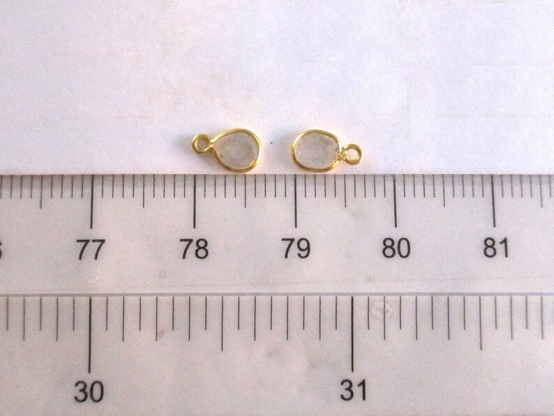 May include: Two gold-toned metal teardrop-shaped pendants with clear glass inserts. The pendants are measured against a ruler, with the top of the pendants aligned with the 78 and 79 markings.