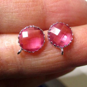 May include: A pair of round, pink gemstone earrings with a faceted surface and a silver-colored, scalloped edge. Each earring has a small loop at the bottom. The earrings are displayed on a light background.