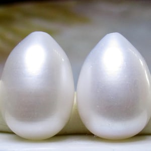 May include: Two pearly white, teardrop-shaped gemstones. The smooth, polished surfaces reflect light, creating a soft, iridescent glow. The gemstones are set against a neutral background, highlighting their elegant form and subtle luster.
