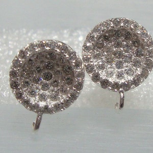 May include: A pair of silver earrings with a round, pave-set diamond design. The earrings have a post and a small, curved hook closure.