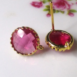 May include: A pair of gold-colored stud earrings with faceted, pink gemstones. The gemstones are set in a gold-colored bezel with a scalloped edge. The earrings are on a white surface with a floral design.