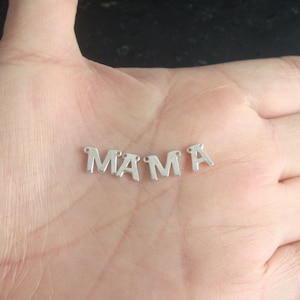 Może przedstawiać: Srebrne metalowe zawieszki z napisem "MAMA". Każda litera ma małą pętlę u góry do nawlekania. Charmsy są wyświetlane na jasnym tle odcienia skóry.
