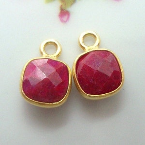 May include: Two gold-toned pendant charms with red gemstone accents. Each charm features a square-shaped gemstone with a faceted design and a small loop at the top.
