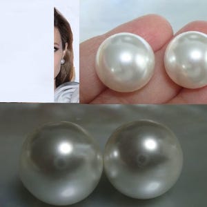 May include: Two large, round, white pearl earrings. The pearls have a lustrous, iridescent sheen. One earring is shown on a person's ear. The other two pearls are shown in a person's hand and on a reflective surface.