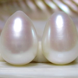 May include: Two lustrous, teardrop-shaped pearls. The pearls have a creamy white base with a subtle pinkish hue at the top, reflecting light. The smooth surface of the pearls creates a soft, elegant appearance.