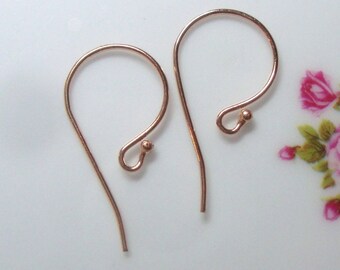 May include: Two copper-colored earring hooks. Each hook has a curved top and a small ball at the end of the curved portion. The hooks are designed for pierced ears and are shown against a soft, blurred background.
