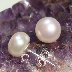 Peut inclure: Une paire de boucles d'oreilles en perles blanches avec des tiges et des dos en argent. Les boucles d'oreilles sont posées sur un fond de cristaux d'améthyste violets.