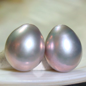 May include: Two iridescent gray pearls with a pearlescent sheen. The pearls are smooth and round, and they are displayed on a white surface.