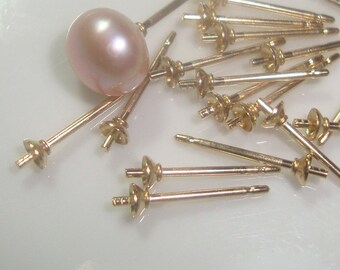 May include: A close-up of a pile of gold-toned earring posts with a single pink pearl lying on the white surface.