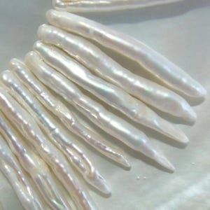 May include: Close-up of a collection of white, elongated pearls. The pearls have a lustrous, iridescent sheen, with subtle variations in color. The shapes are irregular, suggesting a natural origin. The pearls are arranged in a fan-like pattern.