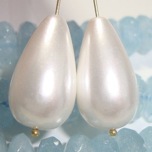 Peut inclure: Deux perles de forme larme de couleur blanc nacré avec des accents dorés, présentées sur un lit de pierres bleu clair.