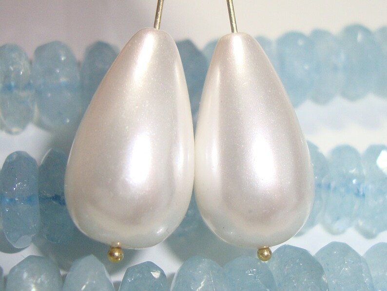 Puede incluir: Dos cuentas de color blanco nacarado en forma de l&aacute;grima con detalles dorados, expuestas sobre un lecho de piedras de color azul claro.