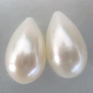 May include: Two pear-shaped, off-white pearls with a lustrous sheen. The teardrop-shaped pearls have a smooth surface and reflect light, suggesting a high-quality finish. The pearls are set against a soft, neutral background.