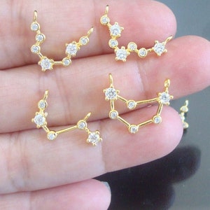 Puede incluir: Cinco dijes de tono dorado con diferentes constelaciones con piedras de circonita cúbica transparentes.