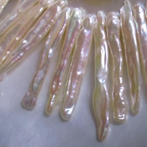 May include: Close-up of a necklace featuring elongated, iridescent pearls. The pearls have a creamy white base with hints of pink and yellow, creating a soft, luminous appearance. The necklace is likely a piece of jewelry.