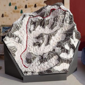 May include: A detailed sculpture of a mountain range, in shades of grey and white, with a red line indicating a route. The sculpture sits on a dark grey angled base. A wooden map is visible in the background.