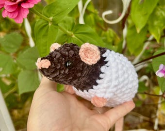 May include: A handmade crochet rat toy in black, white, and peach. The plush rat has a black head, white body, and peach-colored ears and feet. The toy is held in a hand, with a pink flower and green foliage in the background.