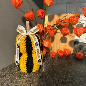May include: A crocheted pumpkin featuring alternating vertical stripes of yellow and black yarn. The pumpkin is topped with a wooden stem and a white ribbon printed with the word Steelers and a football helmet logo tied in a bow at the top.