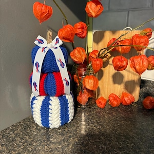 May include: A stacked three-tier crochet pumpkin decoration featuring blue, red, and white yarn segments. The top tier is solid blue, the middle is red and blue, and the bottom is white and blue. A white ribbon with blue buffalo logos is tied at the top.