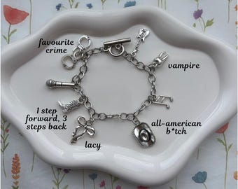 May include: A silver charm bracelet with various charms, including a microphone, cowboy boot, guitar, and a cowboy hat. The bracelet also features charms with the words "favourite crime", "1 step forward, 3 steps back", "lacy", "vampire", and "all-american b*tch".
