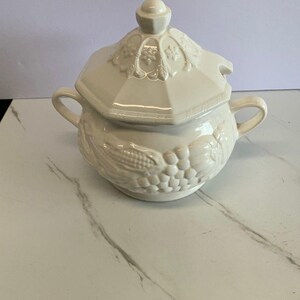 May include: Cream-colored ceramic soup tureen with a lid. The tureen has two handles and is decorated with embossed corn and grape designs. The lid features floral detailing and a small knob.