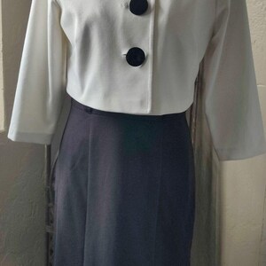 May include: A white cropped jacket with three black buttons and a black skirt. The jacket has a collar and long sleeves.