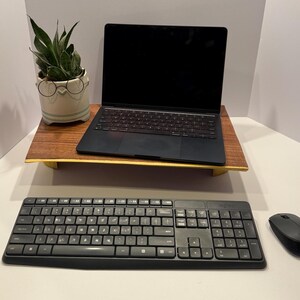 May include: A black laptop computer with a keyboard and trackpad is on a wooden stand. A black wireless keyboard and mouse are on a white surface below the stand. A potted green plant is to the left of the stand.