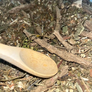 Puede incluir: Una cuchara de madera descansa sobre un montón de hierbas y raíces secas. Las hierbas son una mezcla de verde, marrón y beige, con tallos y hojas visibles. La cuchara es de color claro y está hecha de madera.