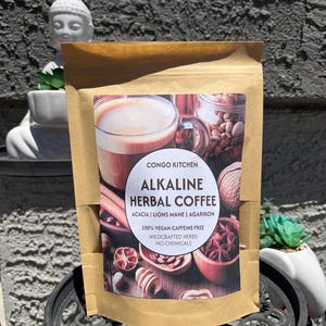 Könnte beinhalten: Ein brauner Papiersack mit Congo Kitchen Alkaline Herbal Coffee. Das Etikett enthält den Text "ACACIA | LIONS MANE | AGARIKON" und "100% VEGAN KOFFEINFREI WILDCRAFTED HERBS OHNE CHEMIKALIEN". Das Bild zeigt eine Tasse Kaffee und verschiedene Kräuter.