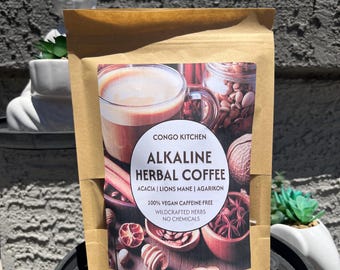 May include: A brown paper bag of Congo Kitchen Alkaline Herbal Coffee. The label includes the text "ACACIA | LIONS MANE | AGARIKON" and "100% VEGAN CAFFEINE-FREE WILDCRAFTED HERBS NO CHEMICALS". The image on the label shows a cup of coffee and various herbs.