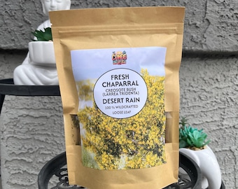 Può includere: Un sacchetto di carta marrone di tè sfuso "Fresh Chaparral". L'etichetta presenta testo e un'immagine gialla e verde della pianta di chaparral. La borsa è esposta su un supporto decorativo in metallo nero, con una statua di Buddha bianca sullo sfondo.