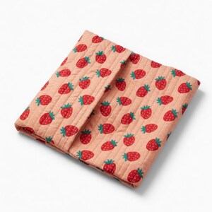 May include: A folded, quilted fabric item with a repeating pattern of red strawberries with green stems on a light peach background. The fabric has a subtle dotted texture. The item appears to be a small blanket or cover.