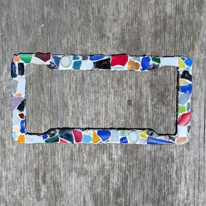 May include: A rectangular license plate frame decorated with a mosaic of multi-colored glass pieces. The frame features a variety of small, irregular shards in shades of red, blue, green, yellow, white, and black, set against a white base.