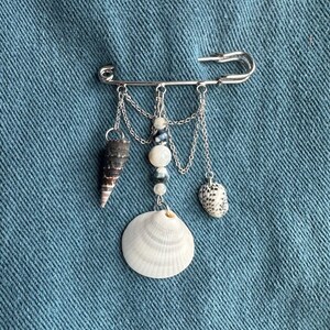 May include: A silver safety pin adorned with dangling seashells and beads. The pin features a variety of shells, including a white scallop shell, a cone-shaped shell, and a speckled shell, all suspended by delicate chains. The background is a textured blue fabric.