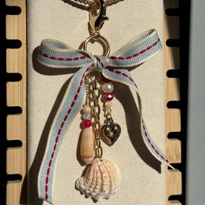 May include: A gold-toned necklace with a blue and red ribbon bow, adorned with a seashell, beads, and a heart charm. The seashell is beige and the bow has red stitching. The necklace is displayed against a neutral background.